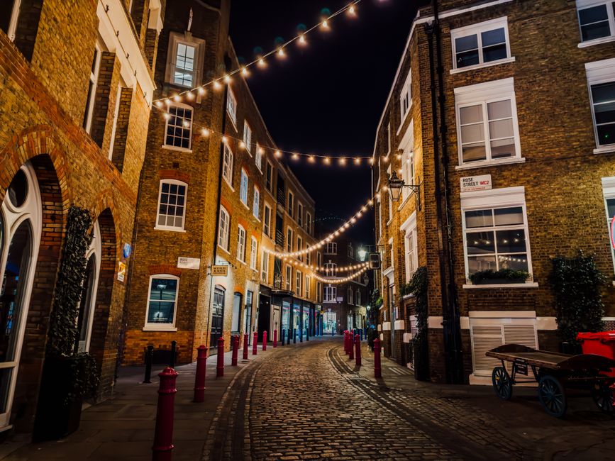 A narrow cobbled street at night illuminated by strings of warm white fairy lights hanging overhead between tall, historic brick buildings on either side. The buildings feature a mix of large, multi-pane sash windows with white frames and some small balconies, with some windows lit from inside. On the right side, a street sign identifies the location as Rose Street WC2. The street is bordered by red bollards along the edge of the pavement, and there are small potted plants and a cart visible on the right side. The scene exudes a quiet, atmospheric ambiance typical of a London alleyway, suitable for illustrating private waste collection or alternative rubbish disposal options in an urban setting. The lighting and textured brickwork contribute to the inviting yet functional environment that Waste Collection Soho might service for rubbish removal.