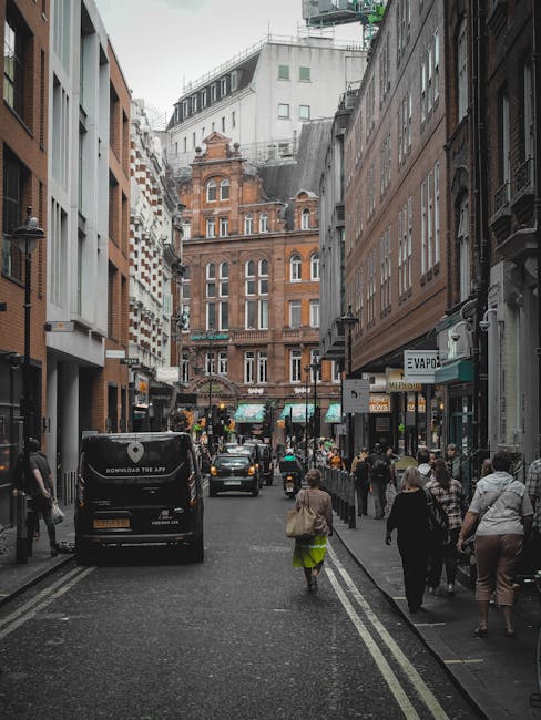 A city street scene viewed from the perspective of someone standing on the pavement. The street is narrow and bordered by tall buildings on both sides, including a prominent red-brick structure with ornate architectural details situated at the end of the street. The buildings feature a mix of modern and historic styles, with large windows and detailed facades. Several pedestrians, some walking alone and others in groups, are moving along the sidewalk on the right, while a variety of vehicles, including a black van and several cars, are parked or driving through the street. Shopfronts with signage, including an air conditioning unit and a storefront with a green awning, line the street. The environment suggests a busy urban area, where private waste collection services such as those offered by Waste Collection Soho may operate in similar alleyways or service streets nearby for rubbish removal and waste management services.