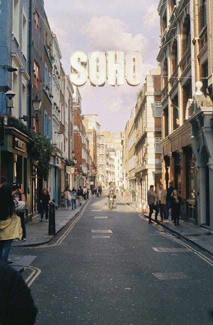 A busy street scene in Soho, London, with pedestrians walking along the pavement on both sides of the narrow roadway. Various shopfronts with classic architectural features line the street, some with outdoor displays and signage. The buildings have a mix of brick and stucco finishes in neutral and pastel tones, with large windows and decorative elements. Overhead, a large, white, three-dimensional sign spelling 'SOHO' is suspended above the street, visible against a partly cloudy sky. The street appears to be in an urban commercial area, with occasional streetlights and bollards present. The scene has natural daylight that highlights the textures and colors of the buildings, pedestrians, and street surface, reflecting typical city centre activity. The environment suggests a vibrant but orderly environment where private waste collection or rubbish removal might answer local needs, with Waste Collection Soho operating in such a context for alternative waste handling.
