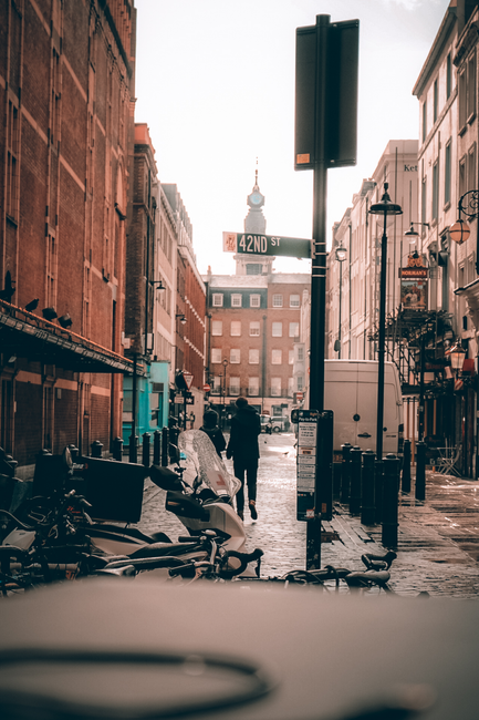 A city street scene viewed from the perspective of someone standing on the pavement. The street is narrow and bordered by tall buildings on both sides, including a prominent red-brick structure with ornate architectural details situated at the end of the street. The buildings feature a mix of modern and historic styles, with large windows and detailed facades. Several pedestrians, some walking alone and others in groups, are moving along the sidewalk on the right, while a variety of vehicles, including a black van and several cars, are parked or driving through the street. Shopfronts with signage, including an air conditioning unit and a storefront with a green awning, line the street. The environment suggests a busy urban area, where private waste collection services such as those offered by Waste Collection Soho may operate in similar alleyways or service streets nearby for rubbish removal and waste management services.