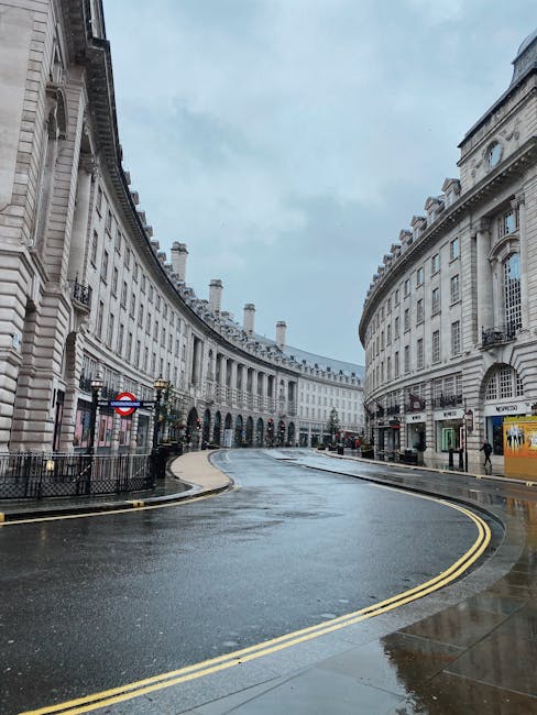 A curved cobblestone street in a city centre features wet pavement reflecting the overcast sky, with classic white stone buildings lining both sides. The buildings have detailed architectural features, including large windows with decorative frames, and some have small balconies with iron railings. On the left, a pedestrian entrance to a Tube station is visible with a London Underground roundel sign, black fencing blocks part of the sidewalk, and traditional street lamps are mounted on the building facades. On the right, a commercial storefront with signage and a yellow advertisement board is partially visible, with a person walking near the entrance. The street appears to be less busy, with no visible vehicles or rubbish, creating a quiet, clean atmosphere that could suggest private waste handling or alternative rubbish collection services in an urban environment like Soho or Piccadilly Circus. The overall scene indicates a typical London cityscape with historic architecture and modern amenities, captured during damp weather conditions.