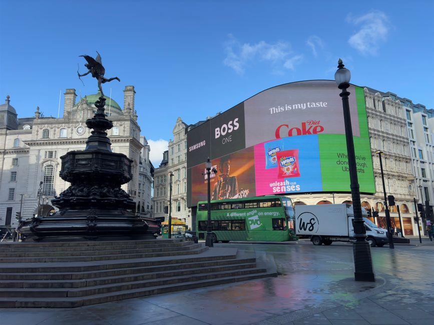 A daytime scene in an urban square featuring a historic dark metal statue of a bird with outstretched wings perched on a pedestal, located on steps leading up to a circular platform. Surrounding the statue are black wrought-iron lamp posts, with a large digital advertising billboard in the background displaying various commercials, including Coca-Cola and Samsung. The billboard curves around the corner of a modern building, with classical architecture visible behind the billboard, characterized by ornate stone facades and decorative spires. A green double-decker bus, a white van with the initials 'WS' on the side, and a small white utility vehicle are parked nearby on a smooth, wet pavement reflecting the scene. The sky above is partly cloudy with patches of blue, and the overall environment appears lively despite the absence of pedestrians, capturing a typical city centre setting suitable for services related to rubbish removal or private waste collection in busy urban areas.